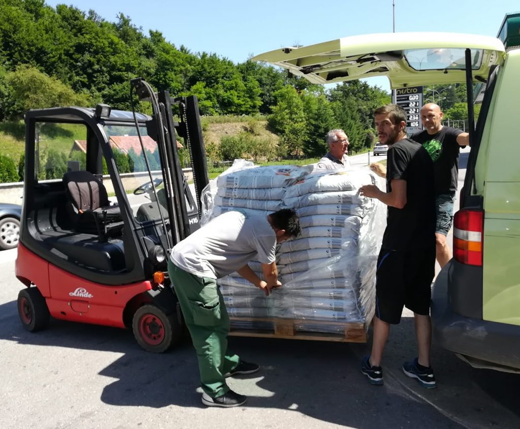 En palletruck står til venstre i bildet og har en pall med store sekker med hundefôr på gaffelen. Det står en mann litt bøyd foran pallen som er i ferd med å kutte opp plasten rundt pallen. Han har grønn langbukse, grå t-skjorte og brunt kort hår. Til høyre for han står det en mann på ca 30 år med sorte shorts og sko, sort t-skjorte og han har mørkt hår med sideskill. Han har kroppen vendt fra oss med hodet vendt til venstre mot kameraet. Til høyre i bildet ses enden av en hvit van som har bakluken oppe. Bak pallen til høyre står det en skallet mann på ca 40 år og lener seg mot vanen.  Rett bak pallen er det en kort eldre mann med grått hår som vi ser fra høyresiden, bare hode og skuldre er synlige bak pallen med hundemat. Bak alle er det en liten grønn ås med løvtrær og taket av et hus med orange taksten skimtes gjennom vinduet på palletrucken.