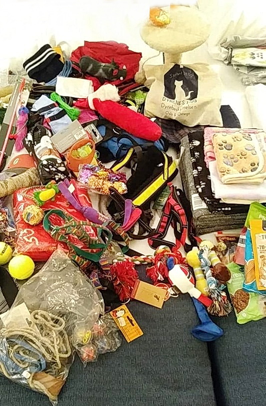 Bildet viser en mengde utstyr  til katter og hunder. Det er mange pledd, tennisballer, hundebånd og tyggeleker. Det er et lite klorestativ til katt, små poser med godis til hunder og et stort handlenett med teksten Dyrenes Hus. Det er et lite berg av gaver.