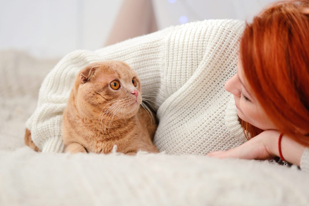 En rødhåret kvinne ligger på et kremefarget pledd og ser på sin røsfargede katt som ser tilbake på henne.